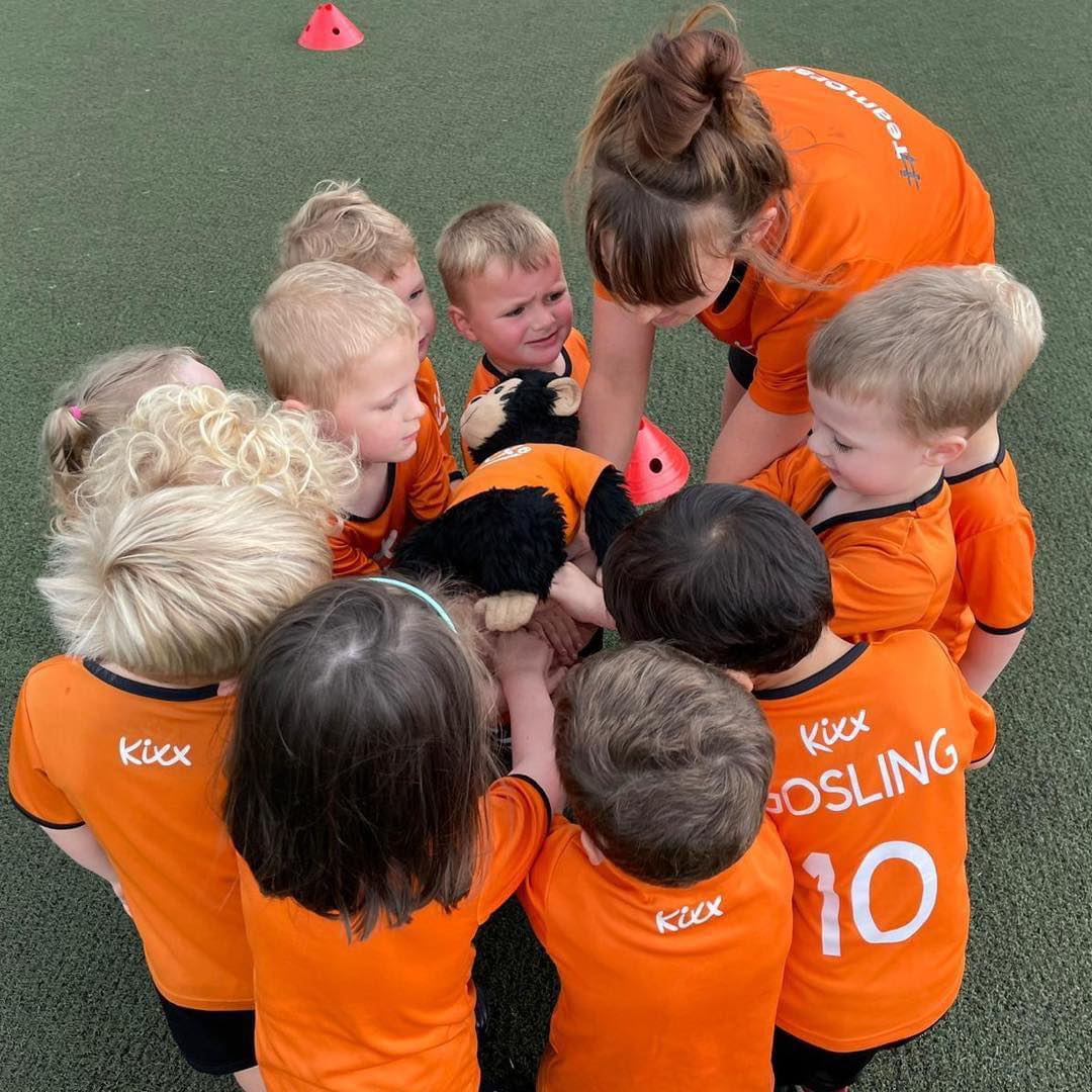 An image of a group of young footballers with their coach, Kerry, doing 'hands in' with a Kixx teddy